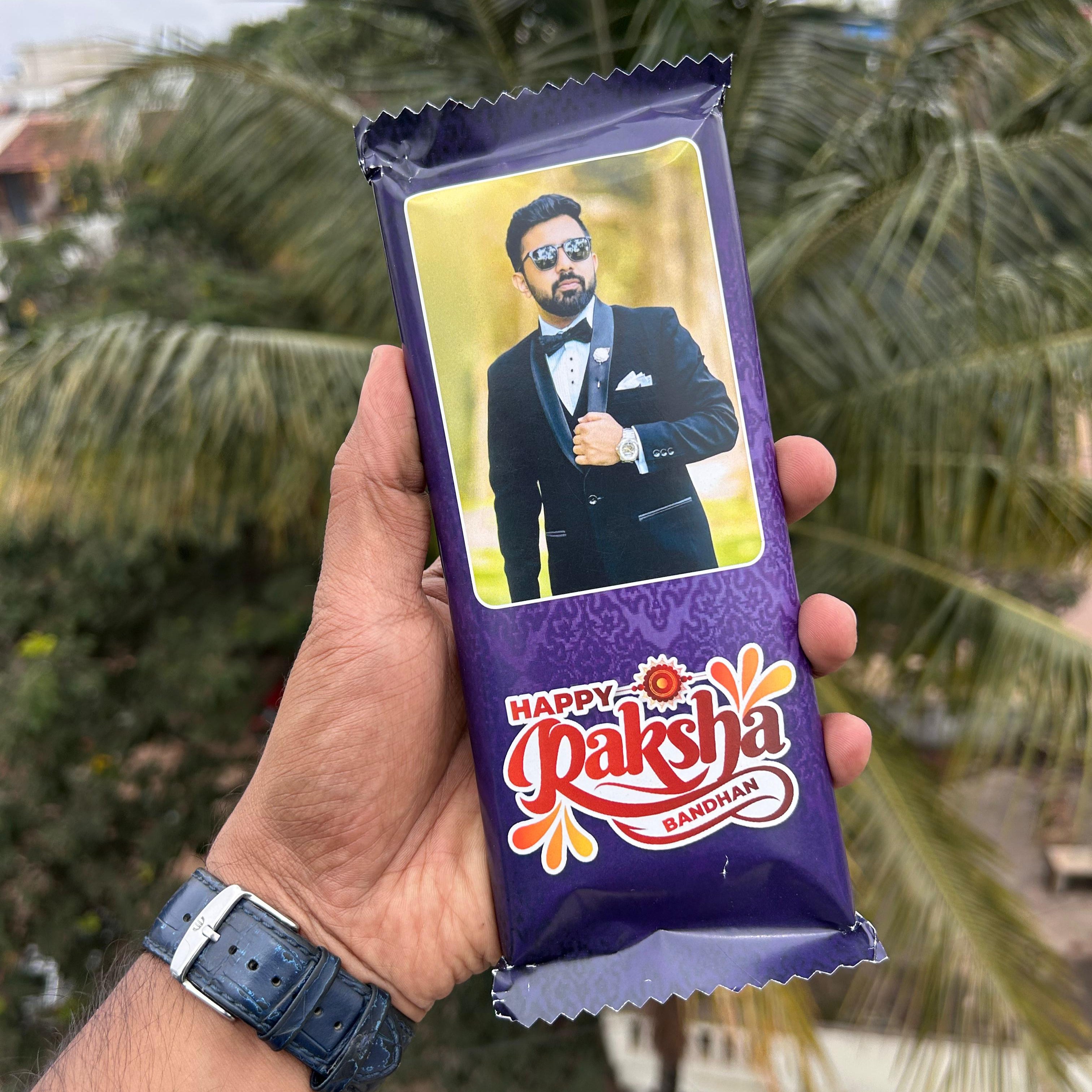 Hand holding a 'Happy Raksha Bandhan' chocolate bar with a person's face on the packaging against a blurred green background. - The Gifting Era